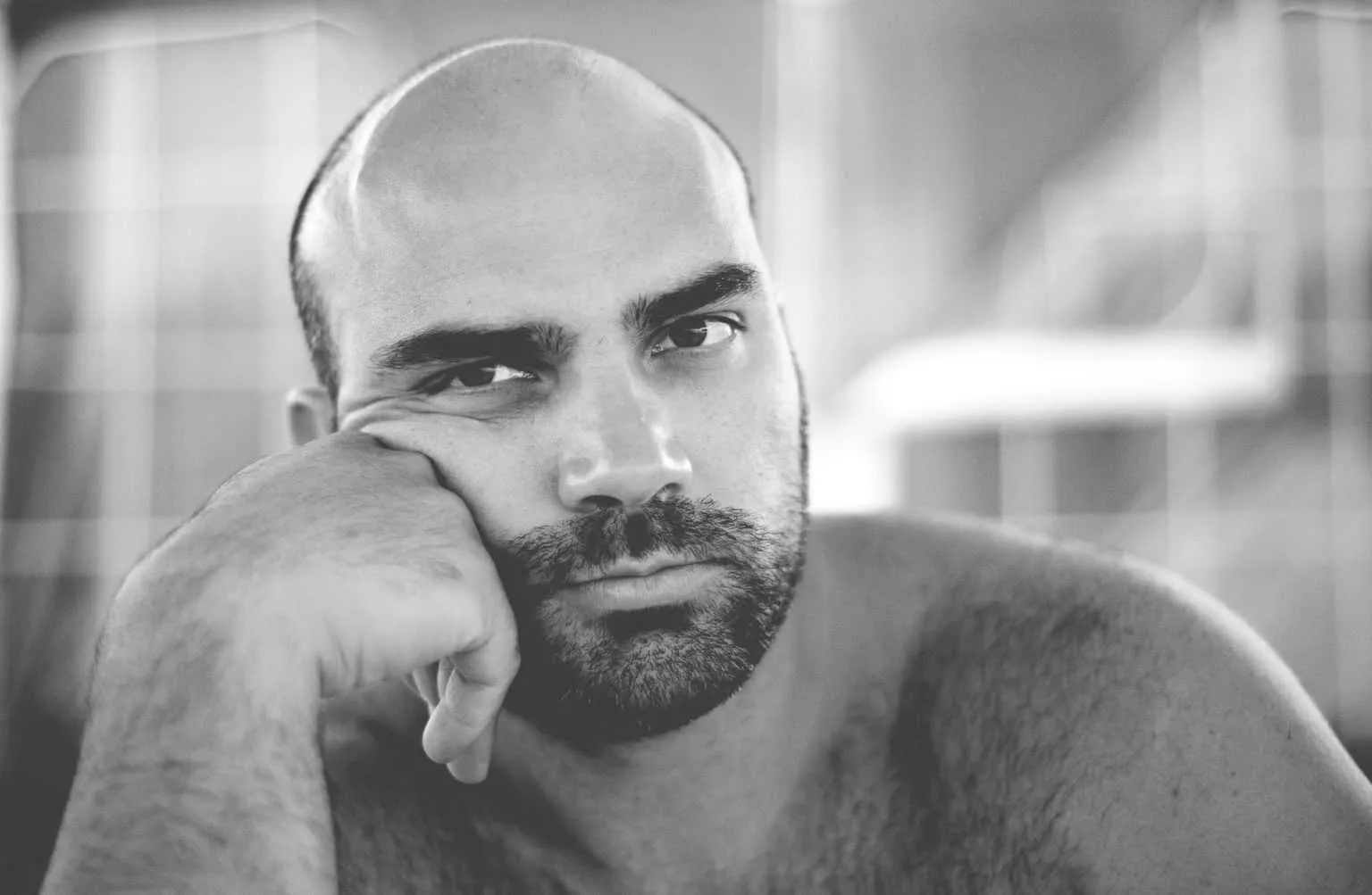Black and white portrait of a bald bearded man resting his head on his hand while looking directly at the camera.
