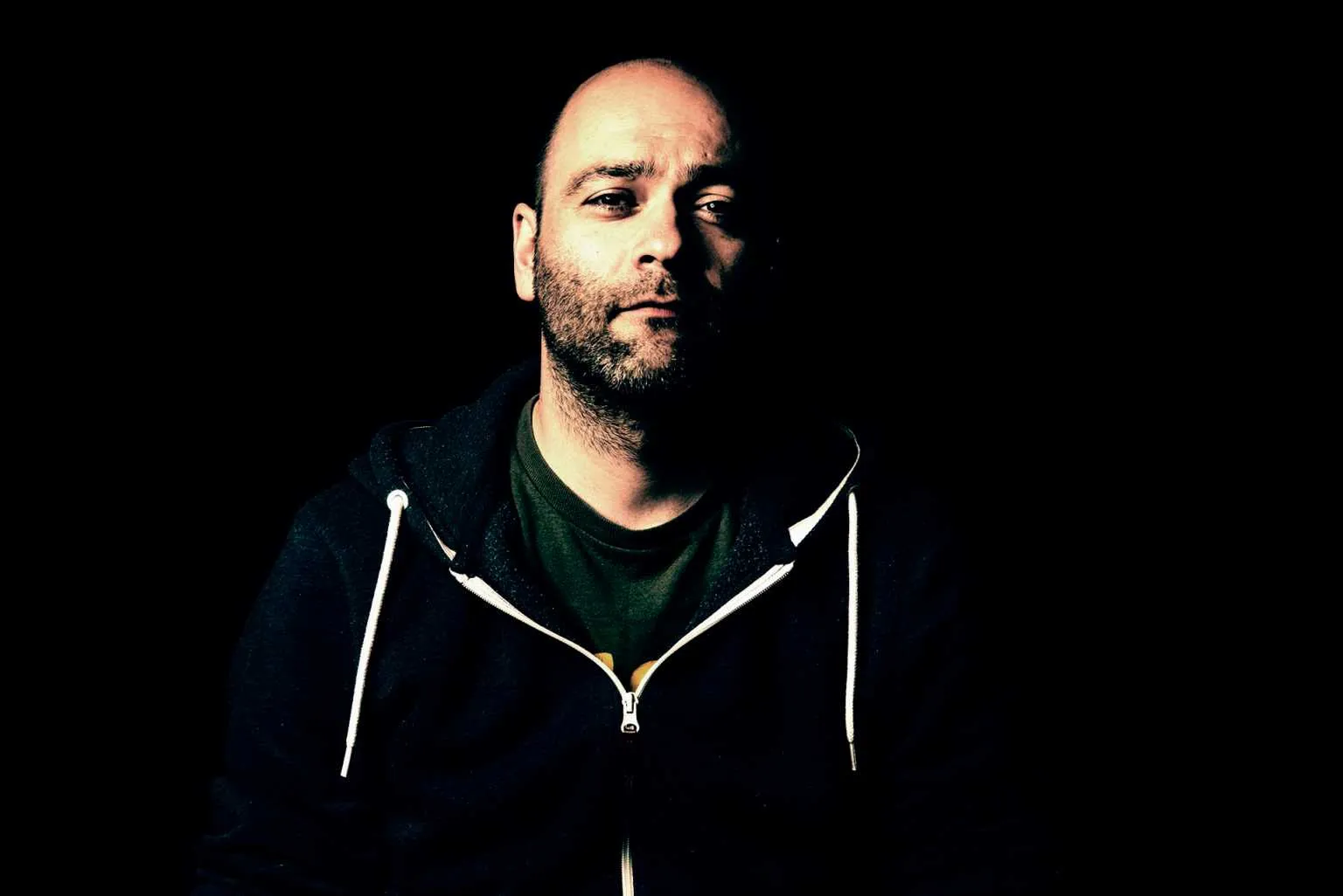 Dramatic low-key portrait of a bald man with a beard wearing a hoodie, half of his face illuminated against a dark background.