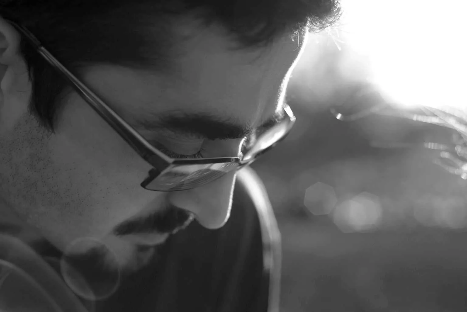 Black and white close-up portrait of a man wearing glasses looking down thoughtfully with soft sunlight and bokeh in the background.