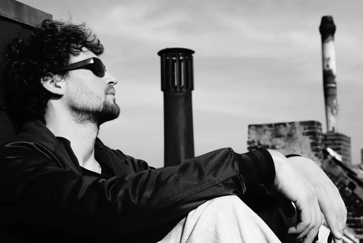 Black and white side profile portrait of a man with curly hair and sunglasses sitting on a rooftop with chimneys in the background.