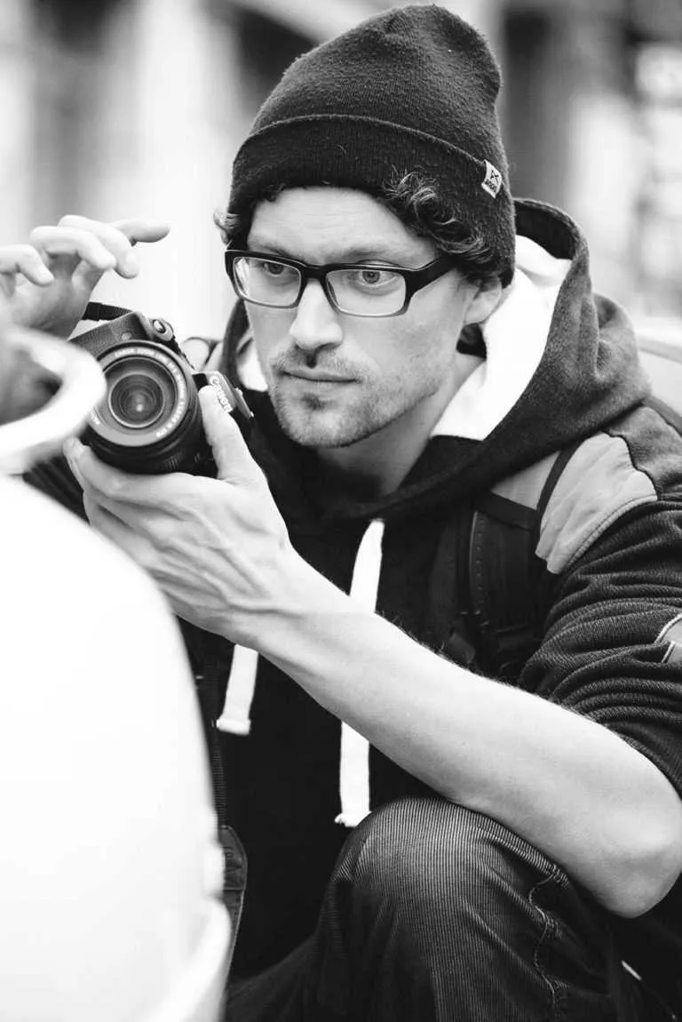 Black and white portrait of a man wearing glasses and a beanie adjusting a camera while photographing someone outdoors.