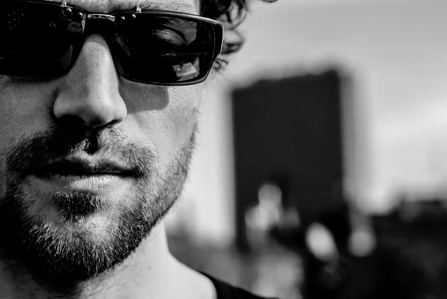 Black and white close-up portrait of a bearded man wearing dark sunglasses with a blurred urban background.