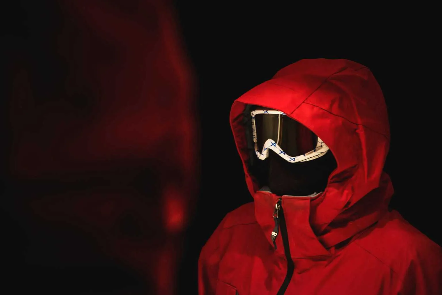 Cinematic portrait of a person wearing a bright red hooded jacket and ski goggles against a dark background.