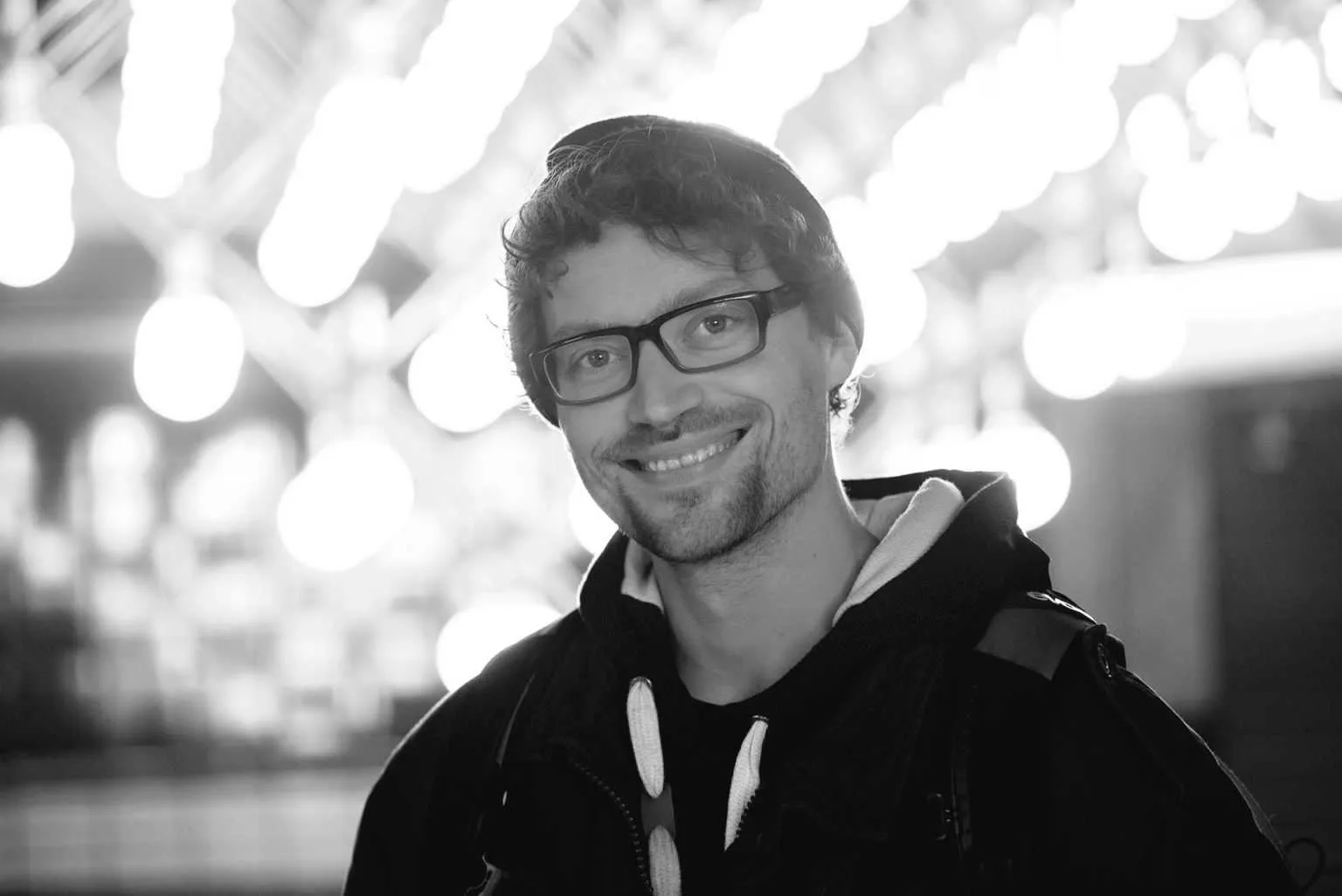 Black and white portrait of a smiling man wearing glasses and a beanie with bright bokeh lights in the background.
