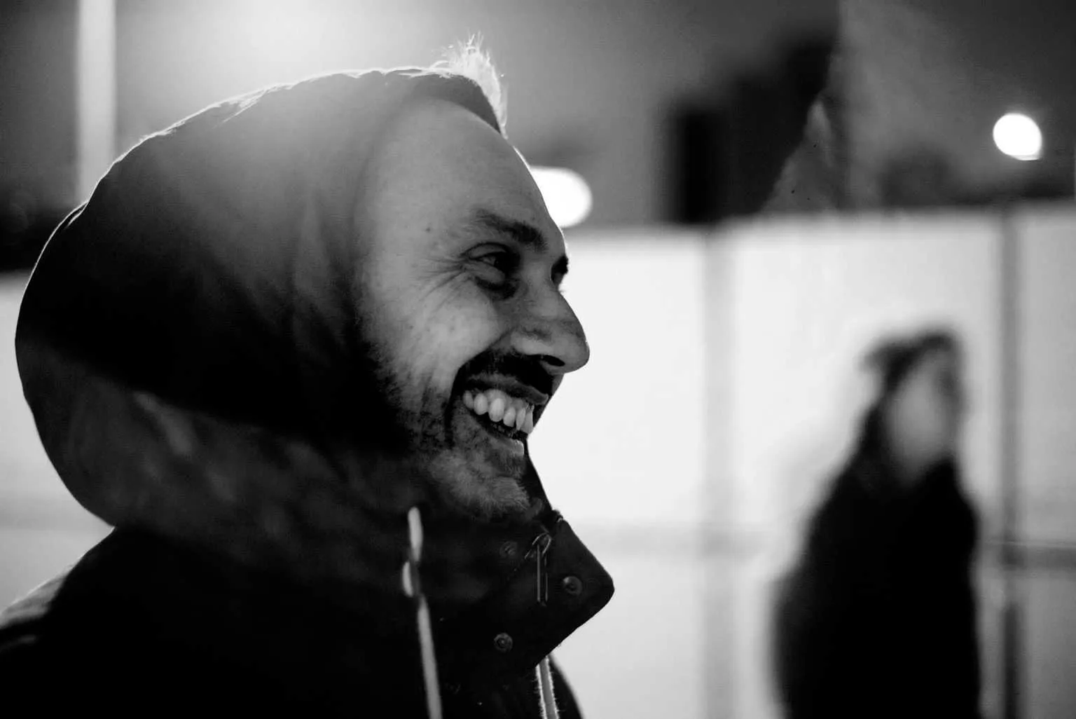 Black and white candid portrait of a smiling man wearing a hooded jacket at night with blurred lights in the background.