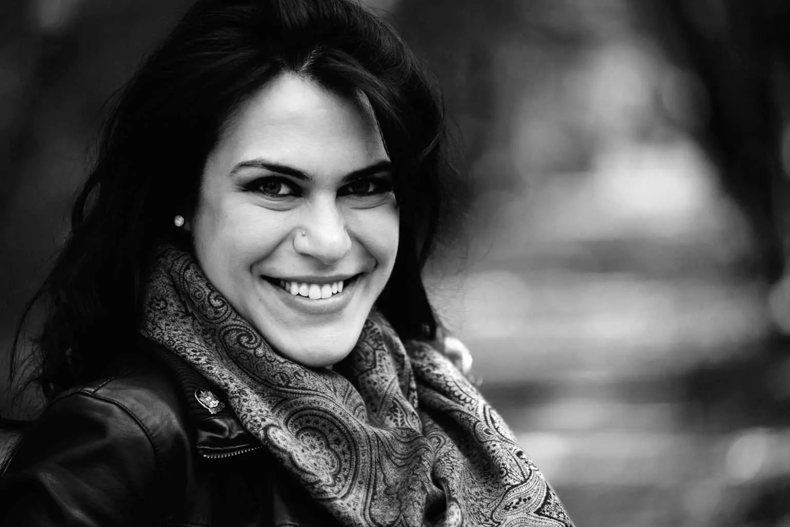 Black and white portrait of a smiling woman with dark hair wearing a patterned scarf and leather jacket outdoors.