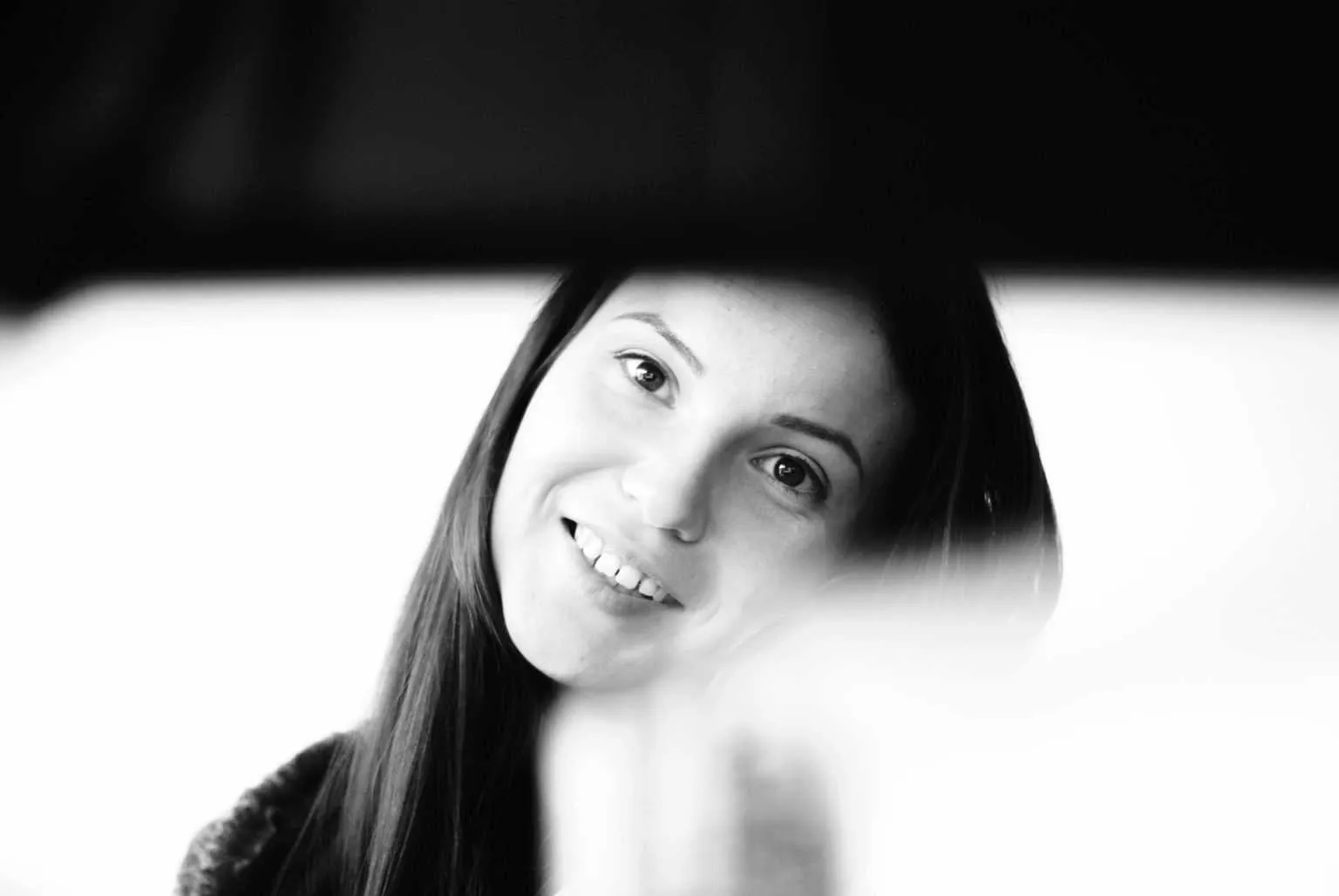 Black and white candid portrait of a smiling woman with long hair looking toward the camera through a soft foreground blur.