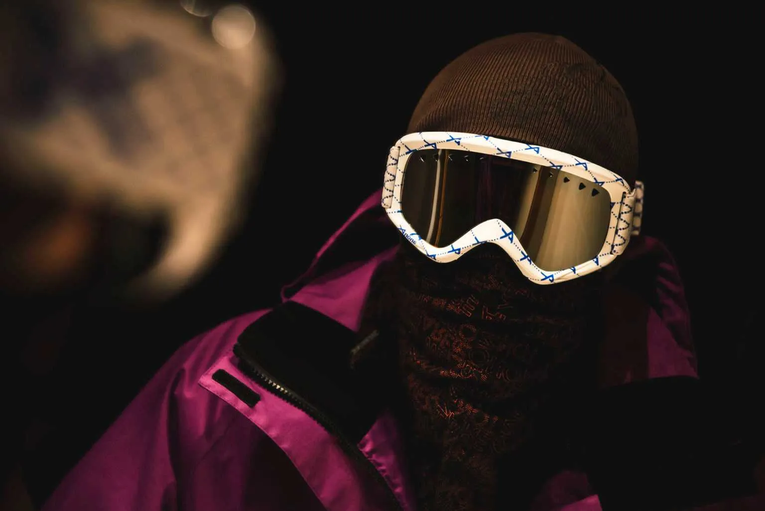 Cinematic portrait of a person wearing snowboard goggles, a beanie, and a face covering with a colorful jacket in dramatic night lighting.