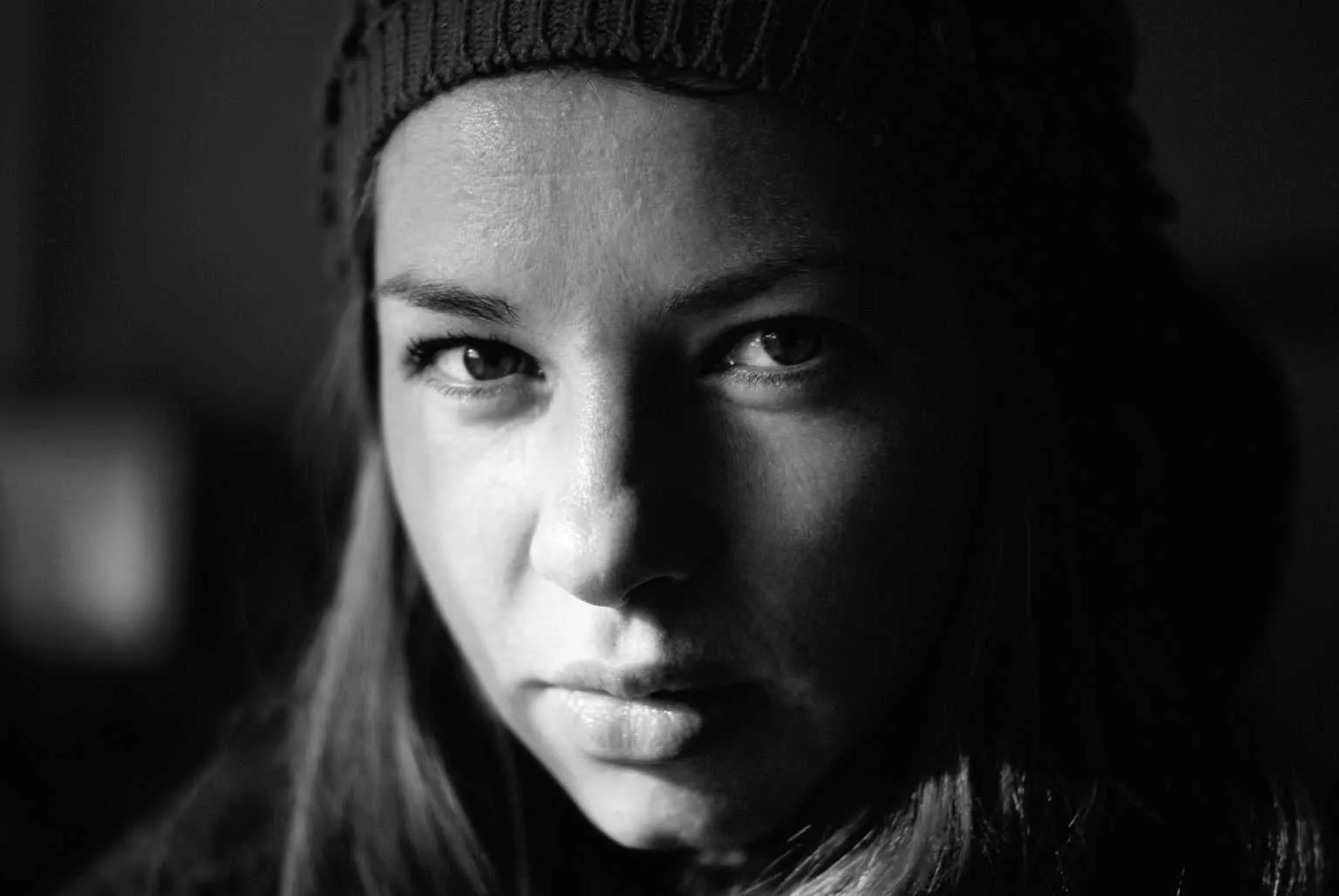 Dramatic black and white close-up portrait of a woman wearing a knit beanie, half of her face illuminated by soft light.