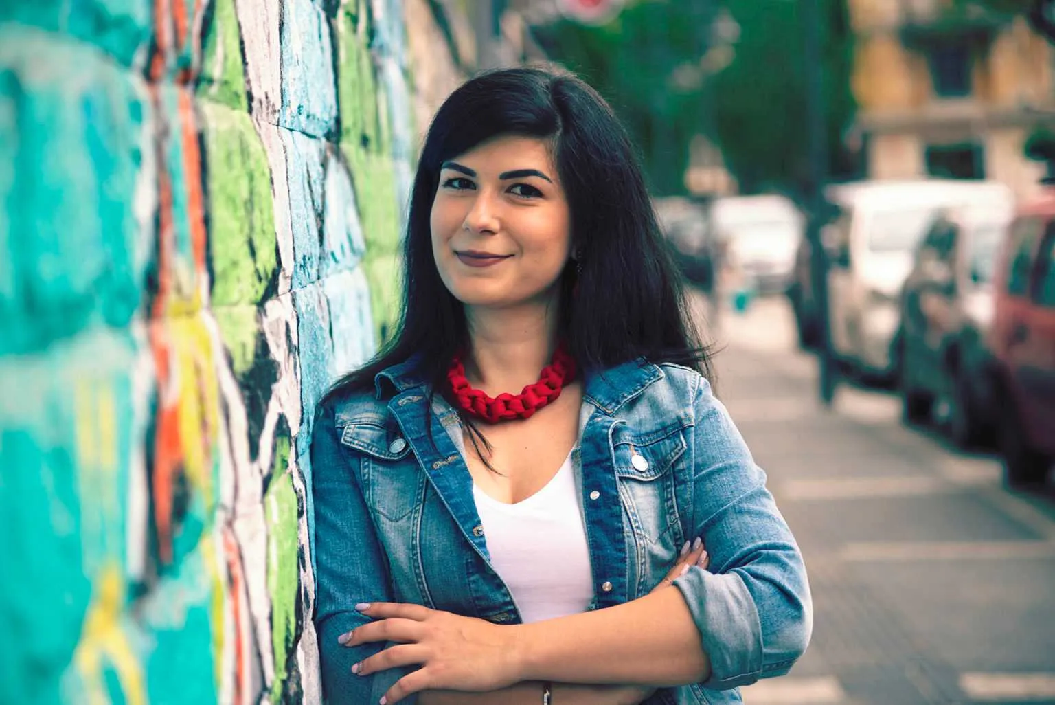 Color portrait of a woman with dark hair leaning against a graffiti-covered wall on a city street.