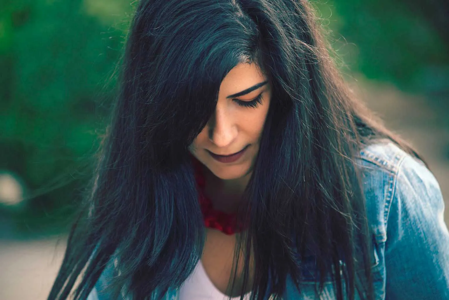 Natural outdoor portrait of a woman with long dark hair looking down, wearing a denim jacket and red necklace.