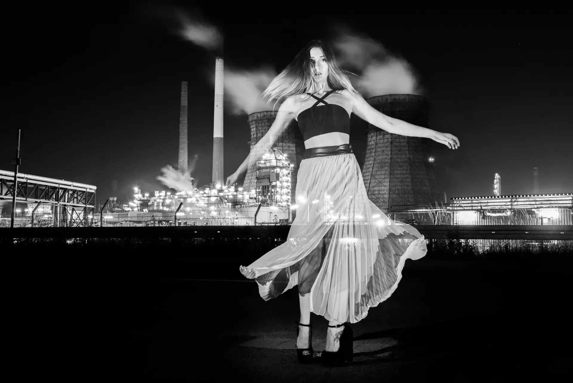 Black and white portrait of a woman dancing in front of illuminated cooling towers and smokestacks at an industrial power plant at night.