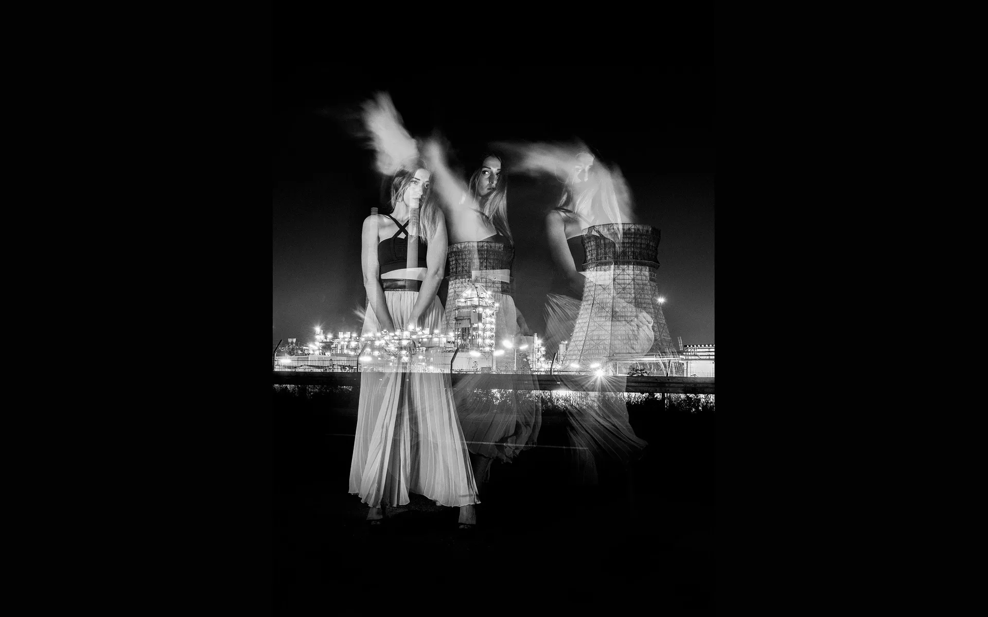 Vertical black and white double exposure of a woman with layered ghosted poses merged with an illuminated industrial power plant at night.