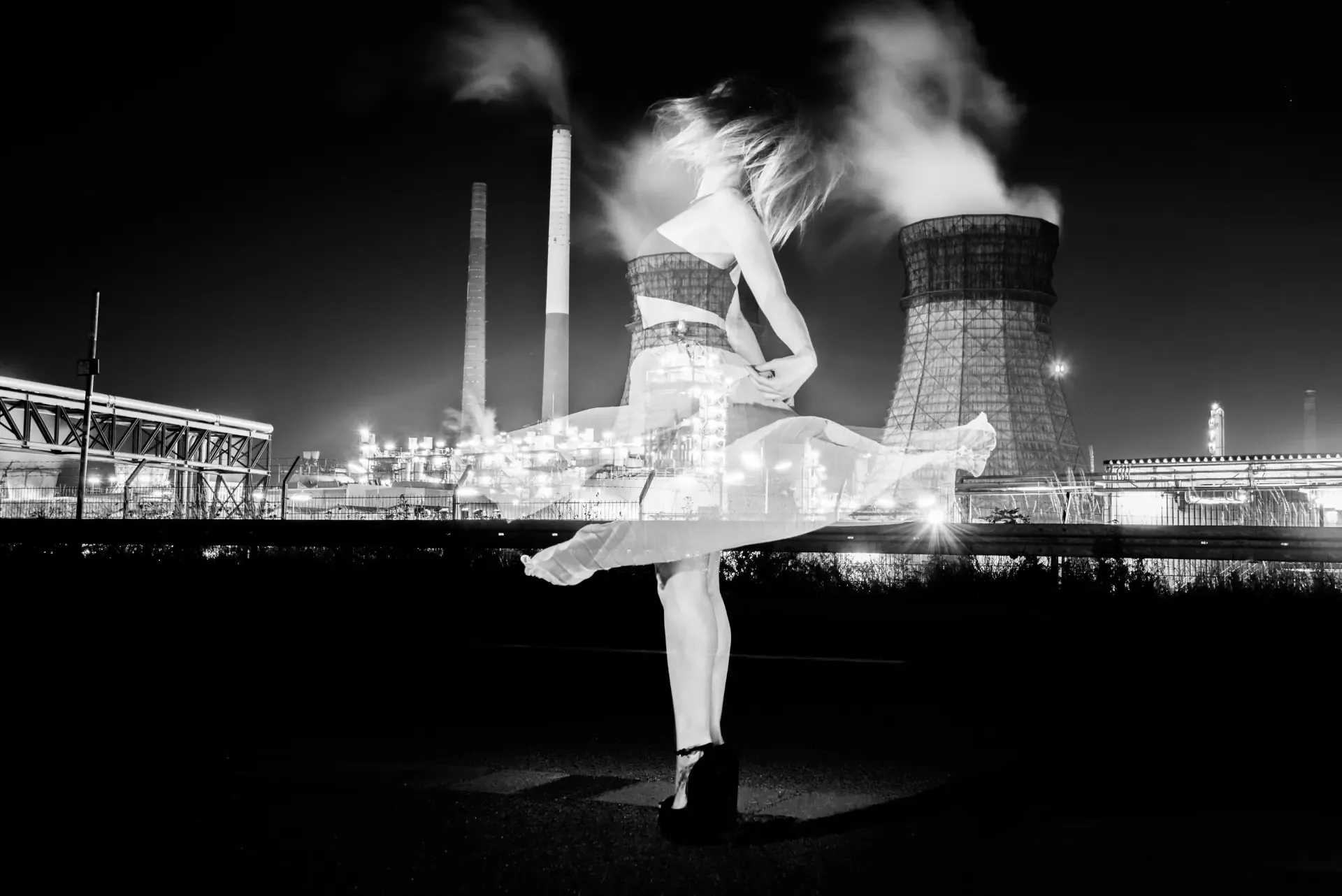 Artistic black and white double exposure showing a spinning dancer layered over a nighttime industrial complex with cooling towers.