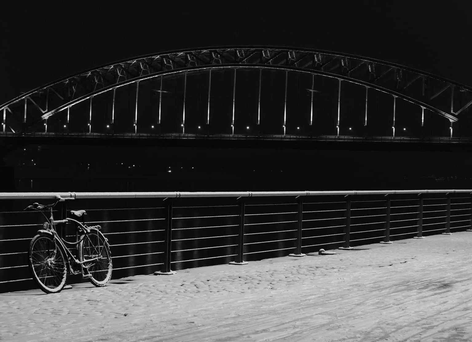 Black and white night photo of an illuminated arch bridge over the rhein in cologne river with a bicycle parked along the riverside railing.