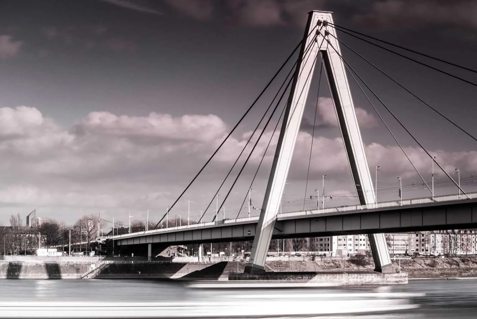 Modern cable-stayed bridge spanning a river with city buildings in the background under a cloudy sky.