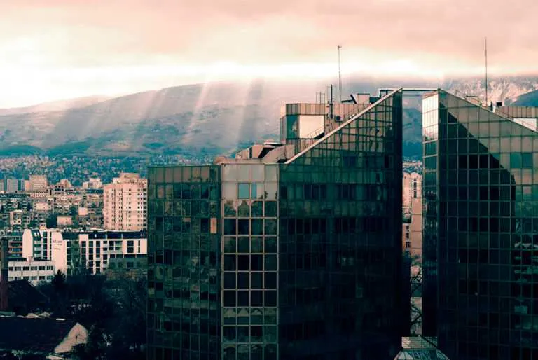 Modern glass office towers with sunlight rays breaking through clouds over a distant cityscape.