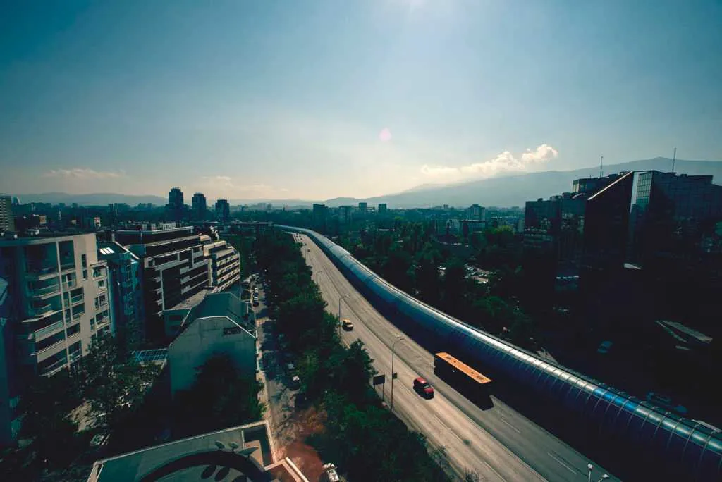 Aerial view of a city highway stretching through modern buildings with mountains in the distance under a bright sky.