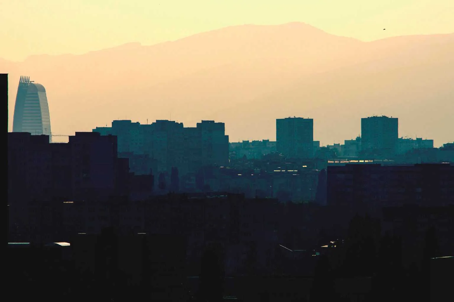 Silhouetted city skyline with apartment blocks and a distinctive modern tower against hazy mountains at sunset.