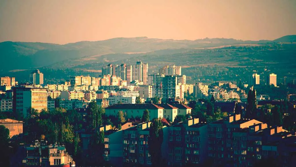 Wide cityscape with residential buildings and towers set against rolling hills in warm evening light.