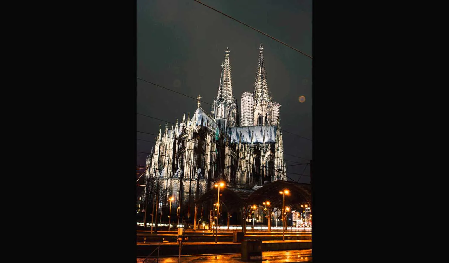 Illuminated Gothic cathedral with twin spires at night, surrounded by city lights and tram lines.