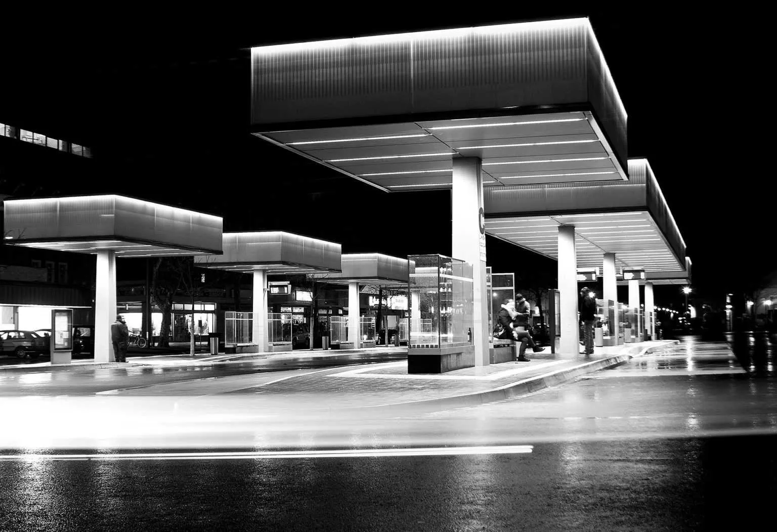 Black and white night photo of a modern bus station with glowing canopy structures and long light trails on wet pavement.