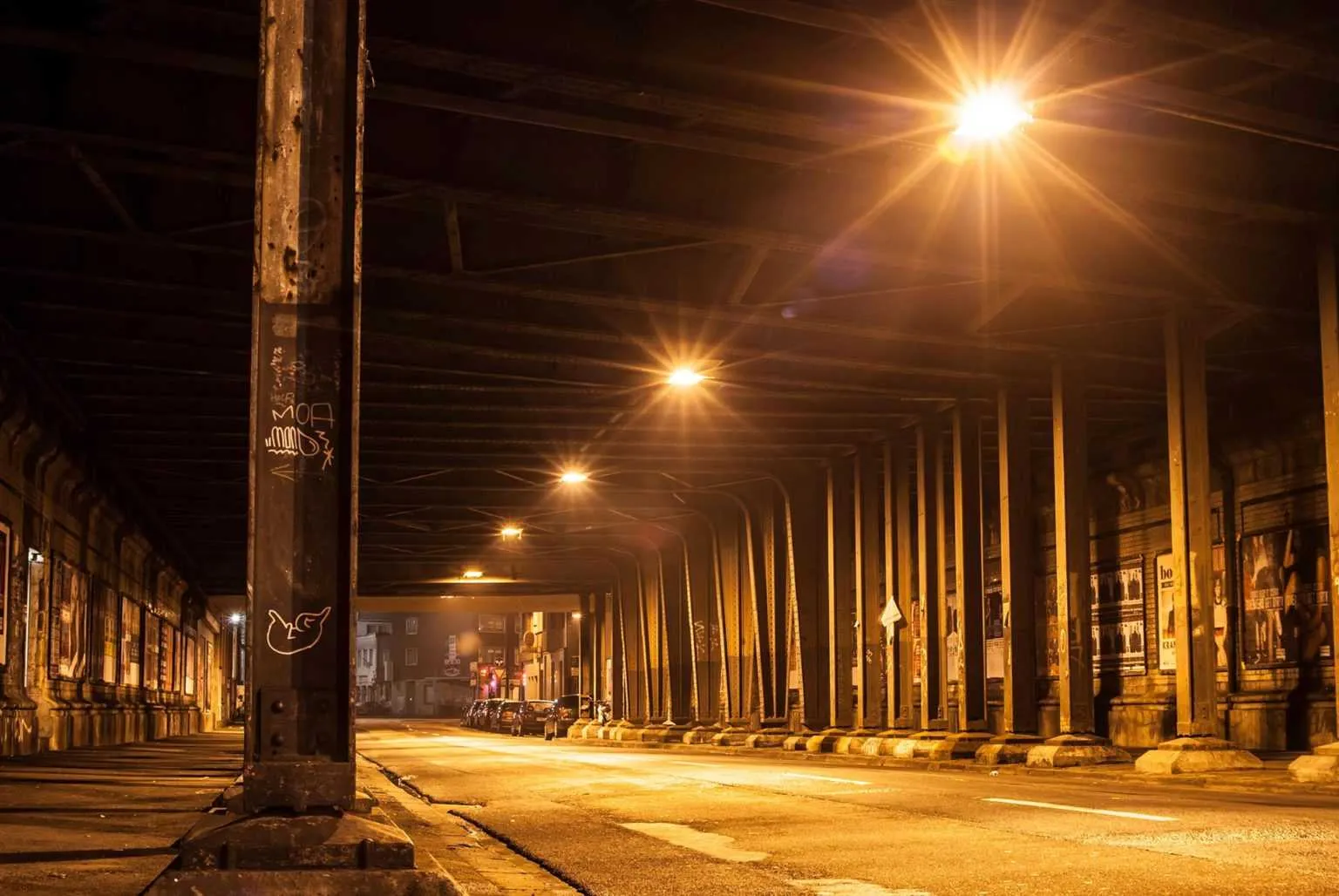 Empty city street running under a steel bridge at night illuminated by warm streetlights.