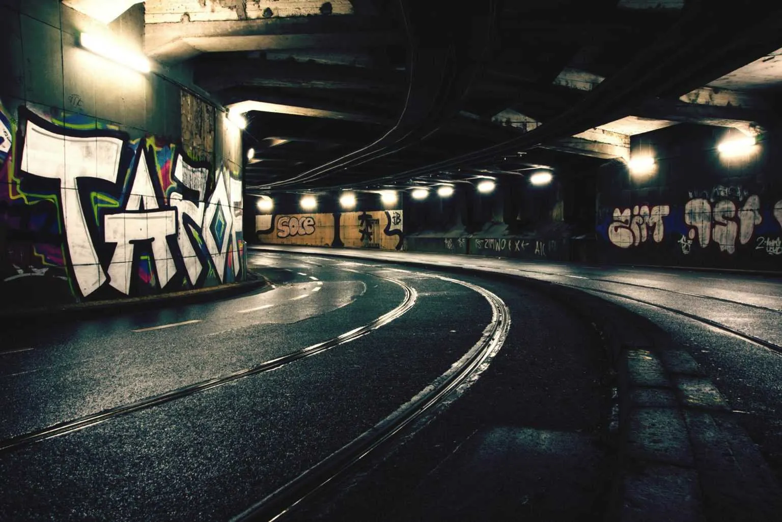 Curving tram tracks running through a dimly lit urban underpass covered with colorful graffiti.